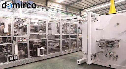 خط تولید اتوماتیک پوشک بهداشتی در حال کار با رول‌های بزرگ مواد اولیه و محصولات نهایی روی نوار نقاله.