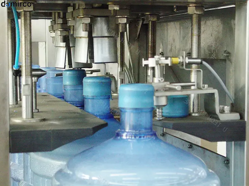 نمای نزدیک از یک دستگاه درب بند گالن صنعتی در حال پلمپ کردن یک گالن پلاستیکی سفید