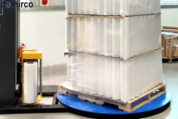 رول‌های فیلم استرچ آبی و شفاف در کنار یک دستگاه پالت پیچ صنعتی در حال کار