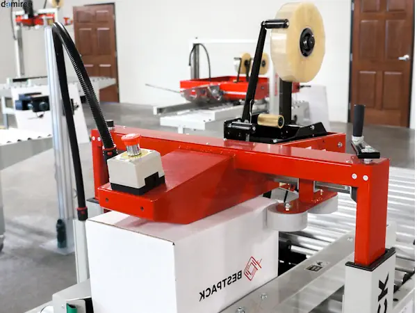 تصویر یک دستگاه چسب زن کارتن کوچک رومیزی در حال چسب زدن یک جعبه قهوه‌ای رنگ