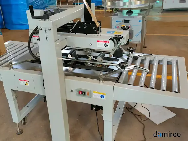 تصویر یک دستگاه چسب زن کارتن صنعتی در حال چسب زدن به یک کارتن در خط تولید