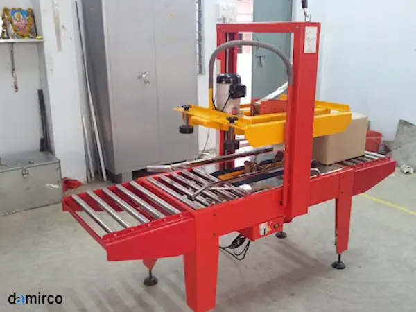 یک دستگاه چسب زن کارتن نیمه اتوماتیک در حال چسب زدن به یک کارتن قهوه‌ای در یک خط تولید صنعتی.